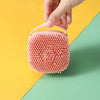 A hand is holding a pink silicone pet bathing massage glove with a textured, bubbly surface against a yellow and green backgr