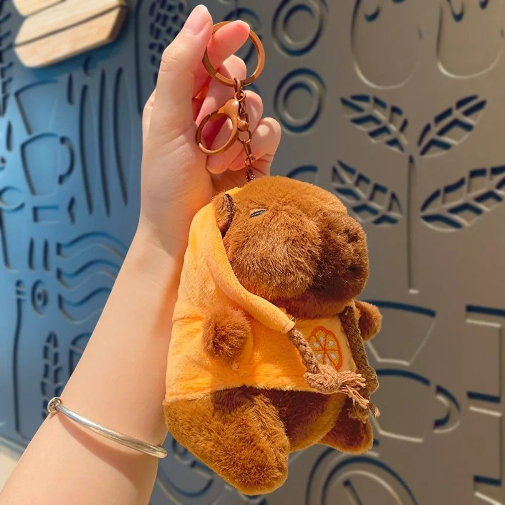 A hand is holding a brown capybara plush doll wearing an orange hoodie and a gold-colored necklace.