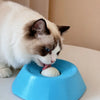 A white cat is interacting with a blue slow feeder bowl, licking a white ball inside.