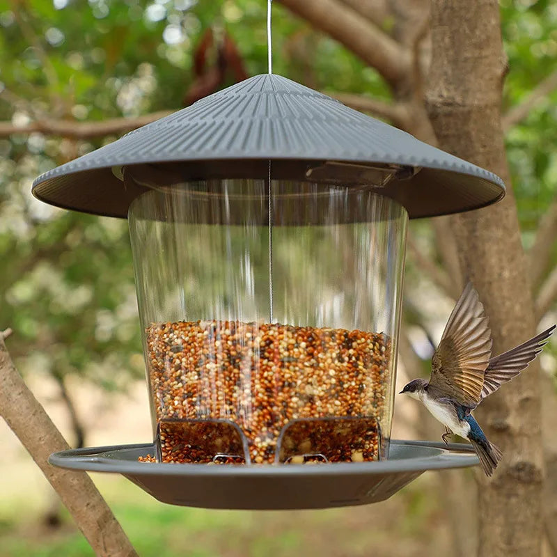 Wasserfester Vogelfutterspender mit konischer Dachform und klarem Glaskörper, von einem Ast aufgehängt.