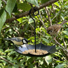 A black umbrella-shaped hanging bird feeder with seeds is suspended from a tree branch, attracting a small bird.