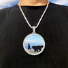 A silver chain necklace features a circular pendant depicting a silhouette of a person and a black bear on a beach.