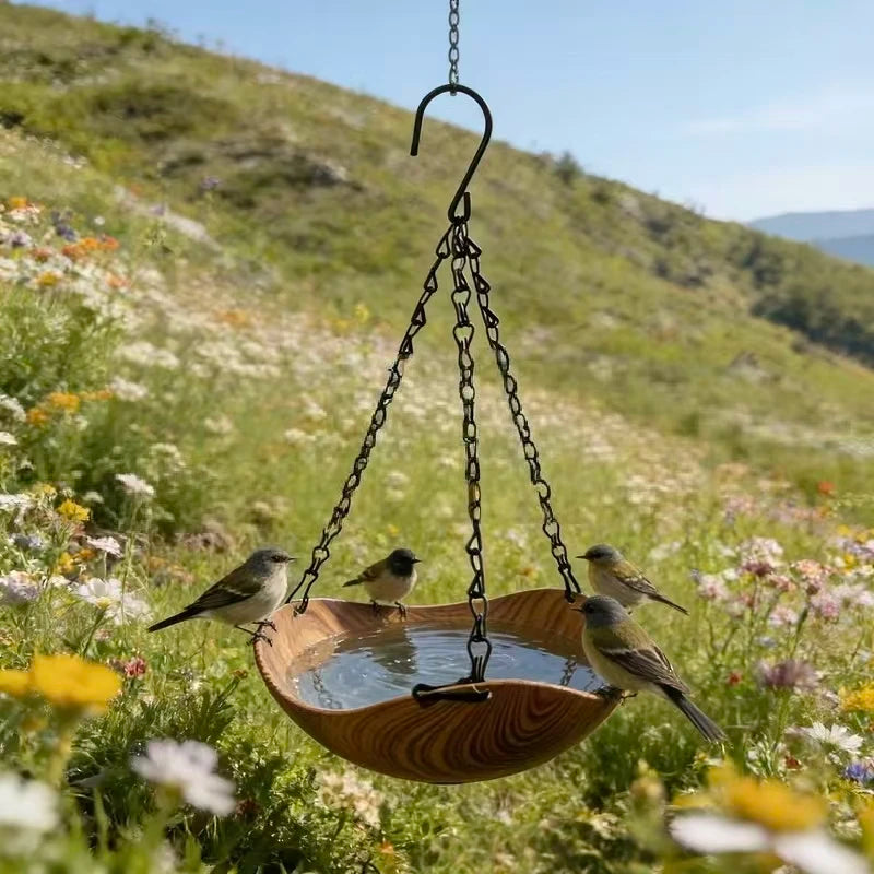 A wooden bird feeder with a bowl is suspended by an iron chain, surrounded by wildflowers on a grassy hillside.