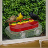 A red suction cup hummingbird feeder with a yellow nectar reservoir and black birds against a green foliage background.