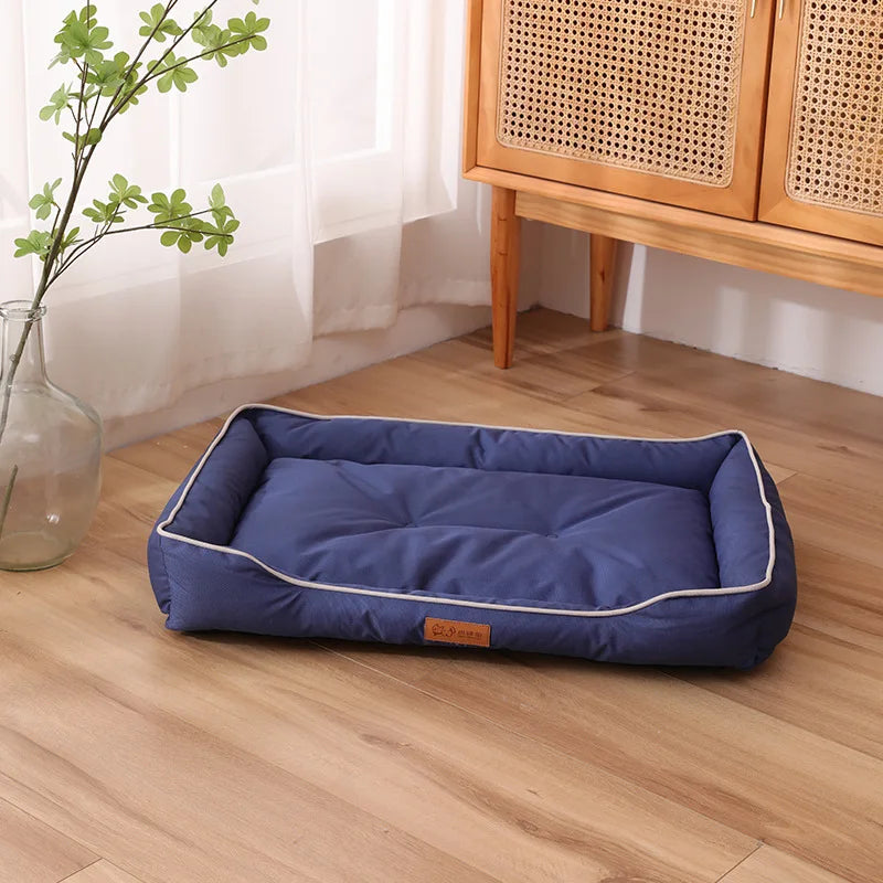 A navy blue waterproof orthopedic dog bed placed on a light wood floor next to a window and wooden furniture.