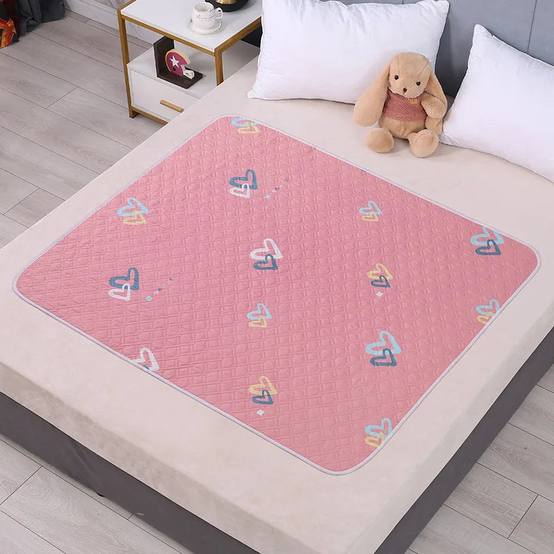 A pink patterned waterproof cartoon bed protector pad is placed on a light-colored table with a white pillow and a stuffed an