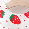 Weißes Stoffkleid mit roter Erdbeere- und kleiner roter Polka-Dot-Muster auf einem Holzbügel.