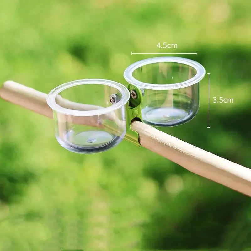 A clear plastic cup with a rim and mounting bracket is attached to a wooden stick, designed as a clip-on bird cage feeder.