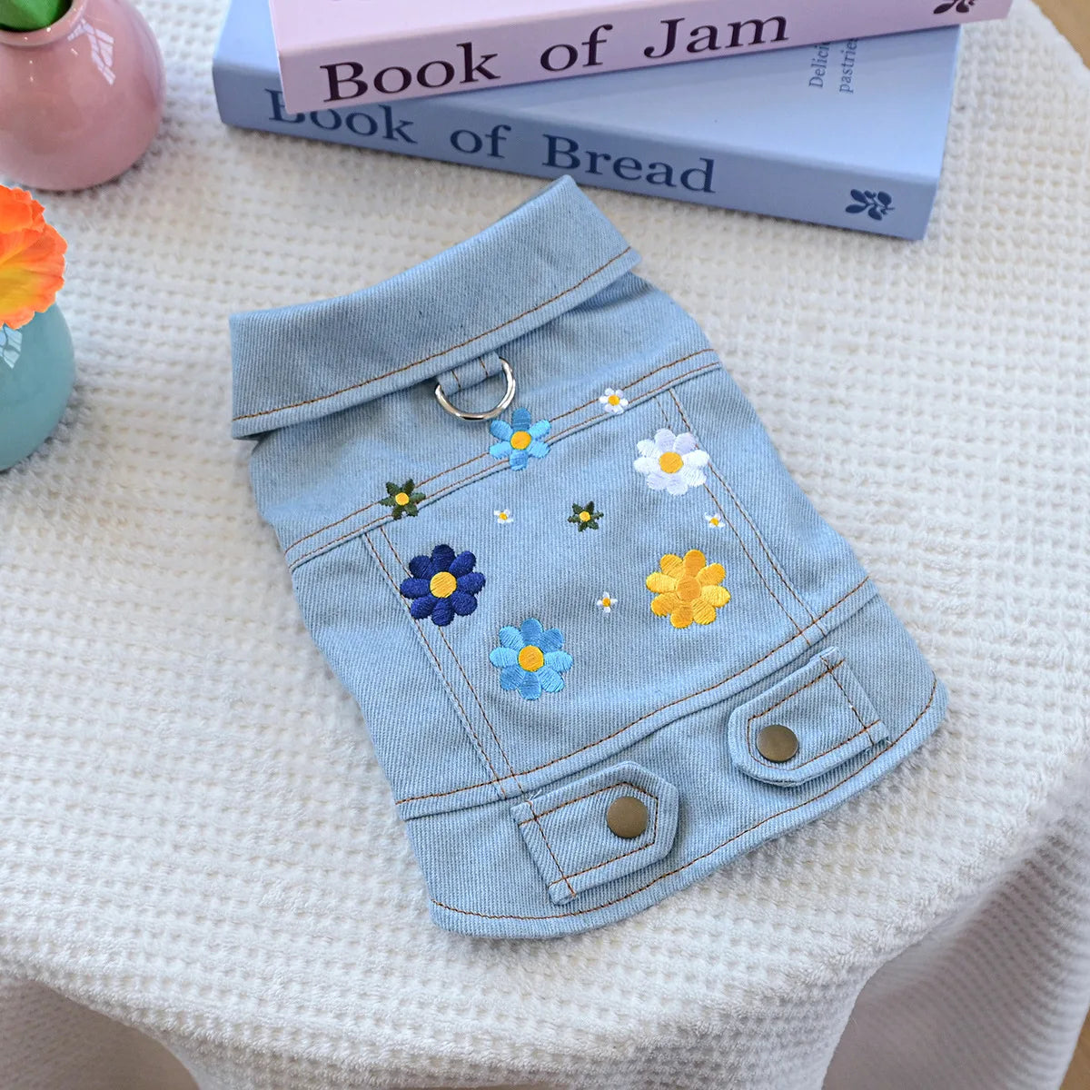 A light blue denim jacket for dogs decorated with colorful fabric flowers and a silver ring detail.