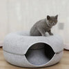 A gray cat is sitting on a round, felt cat bed with a tunnel hideaway for two cats.
