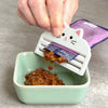A hand is scraping cat food from a purple pouch using a white reusable scraper into a green bowl.