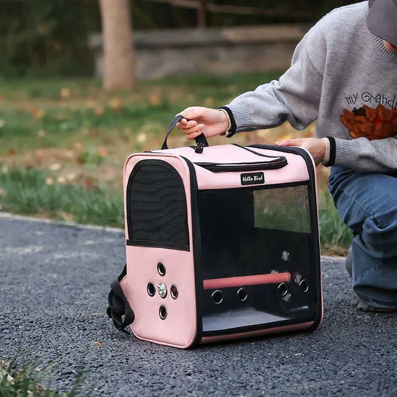 A pink ventilated bird travel backpack with a clear window and feeding cups is being held by a person outdoors.