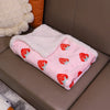 A pink pet blanket with a white sherpa lining and red strawberry floral patterns is resting on a brown leather sofa.