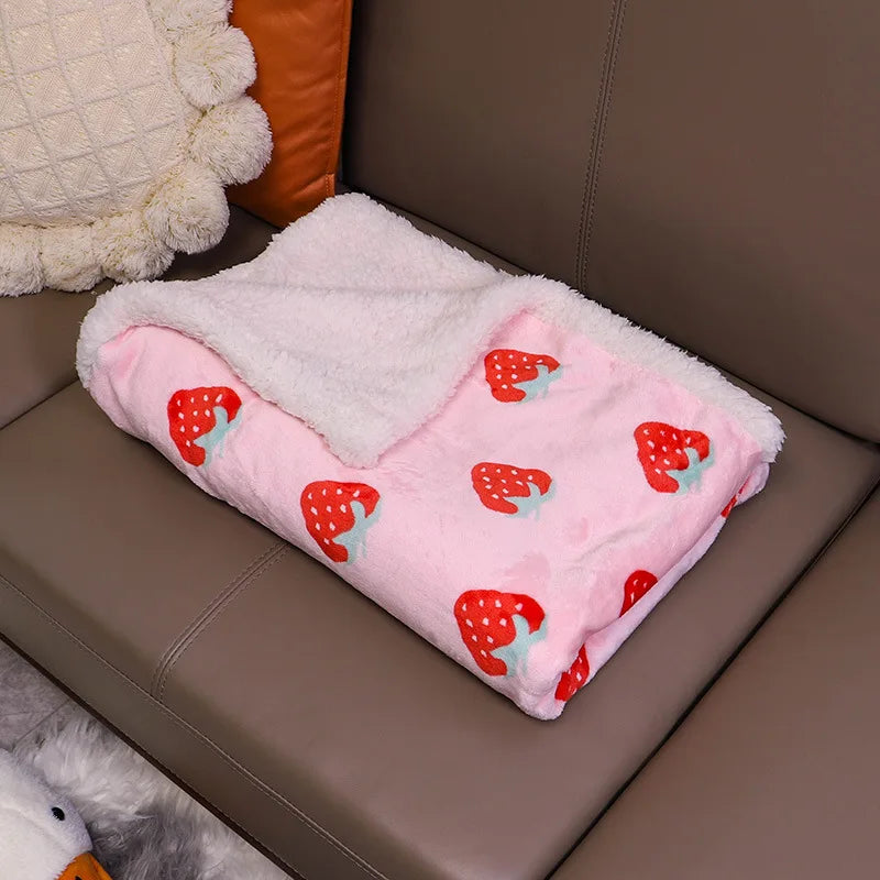 A pink pet blanket with a white sherpa lining and red strawberry floral patterns is resting on a brown leather sofa.