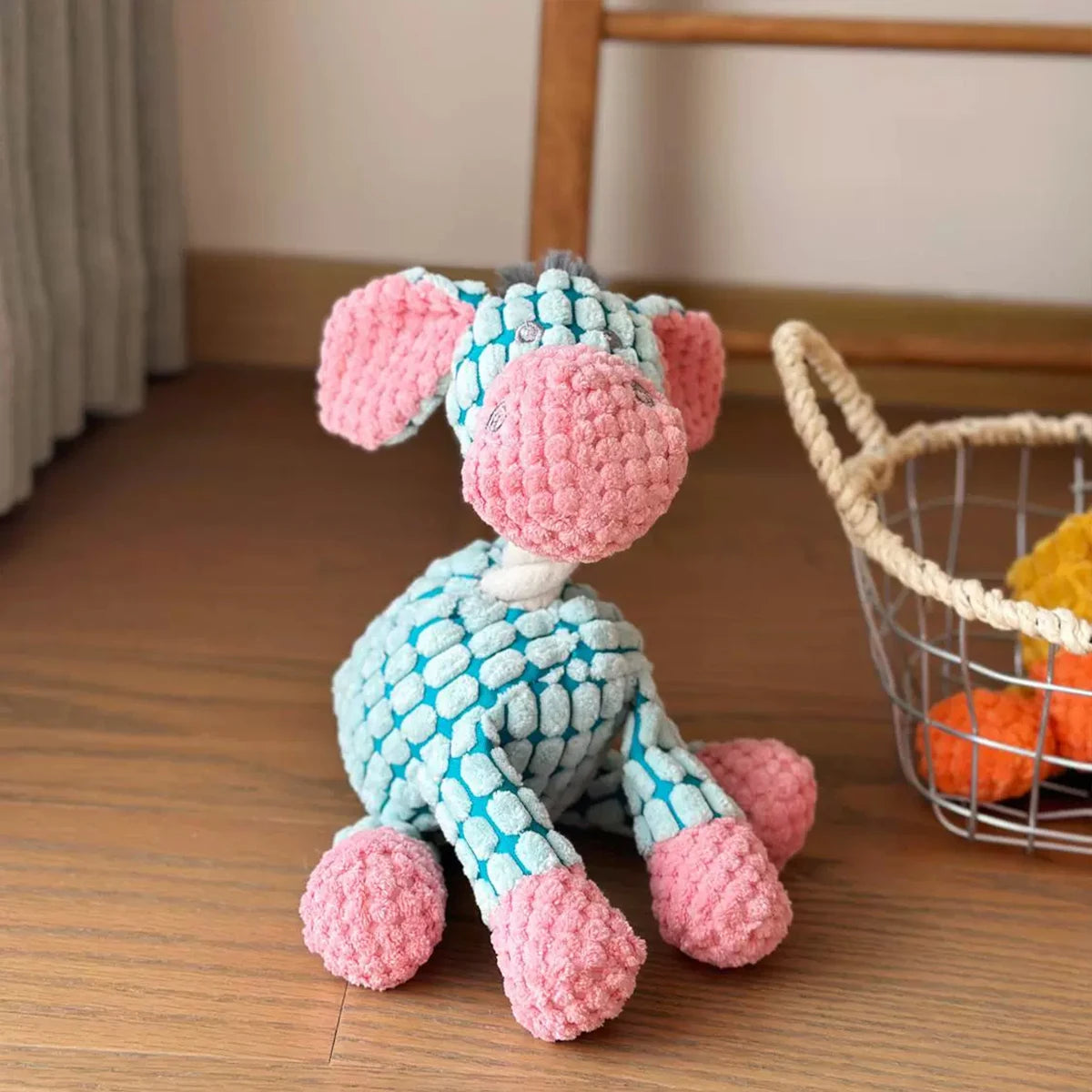 A handmade plush toy of a donkey with pink and blue fur sitting next to a wire basket.
