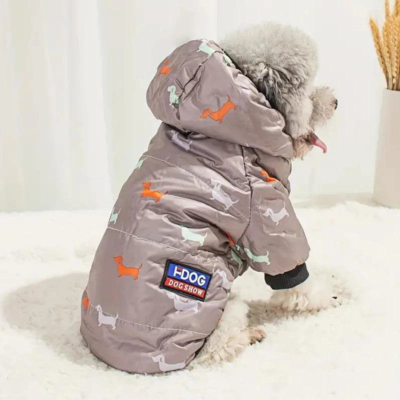 Un perro gris esponjoso vistiendo una chaqueta acolchada con capucha de patrones naranjas y verdes, tumbado sobre una superfi
