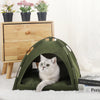 A green cozy pet tent bed for cats is shown resting on a white surface next to potted plants and a wooden drawer.