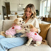 A woman is holding two golden retriever puppies wearing pink strawberry embroidered dog dresses on a couch.