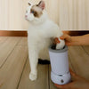 A white and brown cat is being cleaned by a hand using an automatic pet paw cleaner cup.
