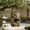 A carved wooden owl feeder made from a tree trunk hangs from a pine branch in an outdoor garden setting.