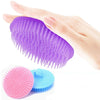 A hand holds a purple silicone hair and scalp massage brush next to a pink and blue silicone brush.