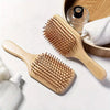 A wooden bamboo hair brush with massage bristles is shown next to a wooden bowl and a white towel.