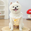 A fluffy white dog wearing a light yellow cotton vest with a lace collar and cartoon print, sitting on a wooden mat.