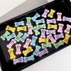 A collection of bone-shaped hair clips for pet grooming in various pastel colors arranged in a black tray.