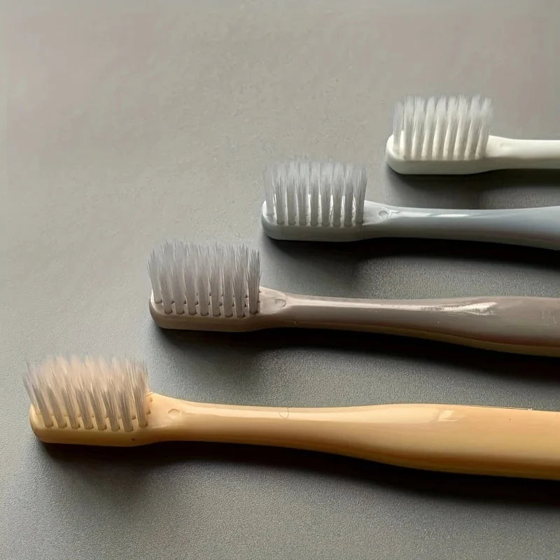 A collection of four soft-bristle travel toothbrushes in various shades of white and beige resting on a gray surface.