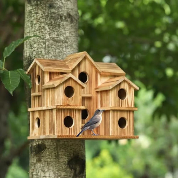 A handmade wooden bird house with a pitched roof and various sized holes is mounted on a tree trunk.