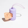 A purple portable dog poop bag dispenser with a pink strap is shown next to a crumpled piece of paper.