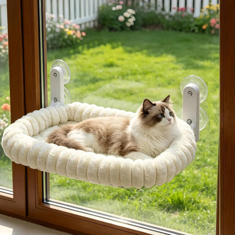 A fluffy tabby cat is relaxing in a white, cushioned hammock on a window ledge, secured by suction cup hooks.