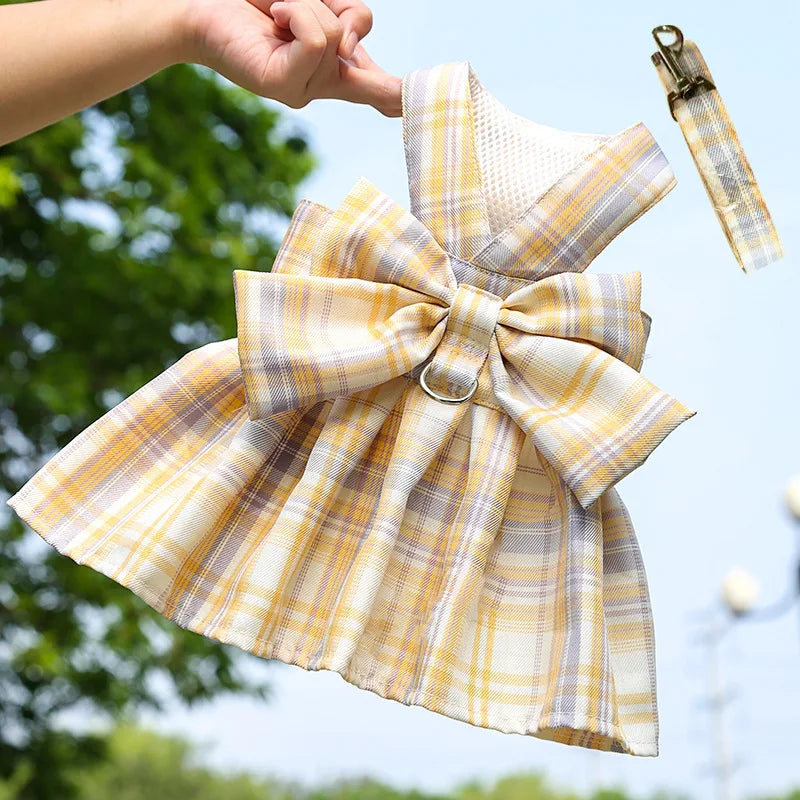 A dog wearing a yellow and beige plaid dress-up outfit with a bow and harness is being held in the air.