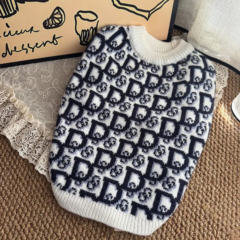 A white and navy knit sweater for dogs with a repeating diamond pattern, displayed on a brown surface.