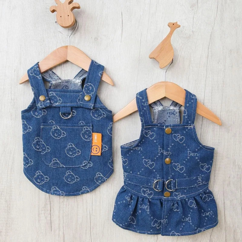 A denim dog harness dress with a D-ring is displayed on a wooden hanger against a light background.