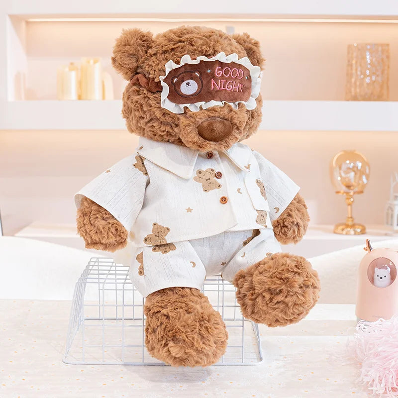 A brown teddy bear plush wearing a white pajama top and pants with a decorative eye mask