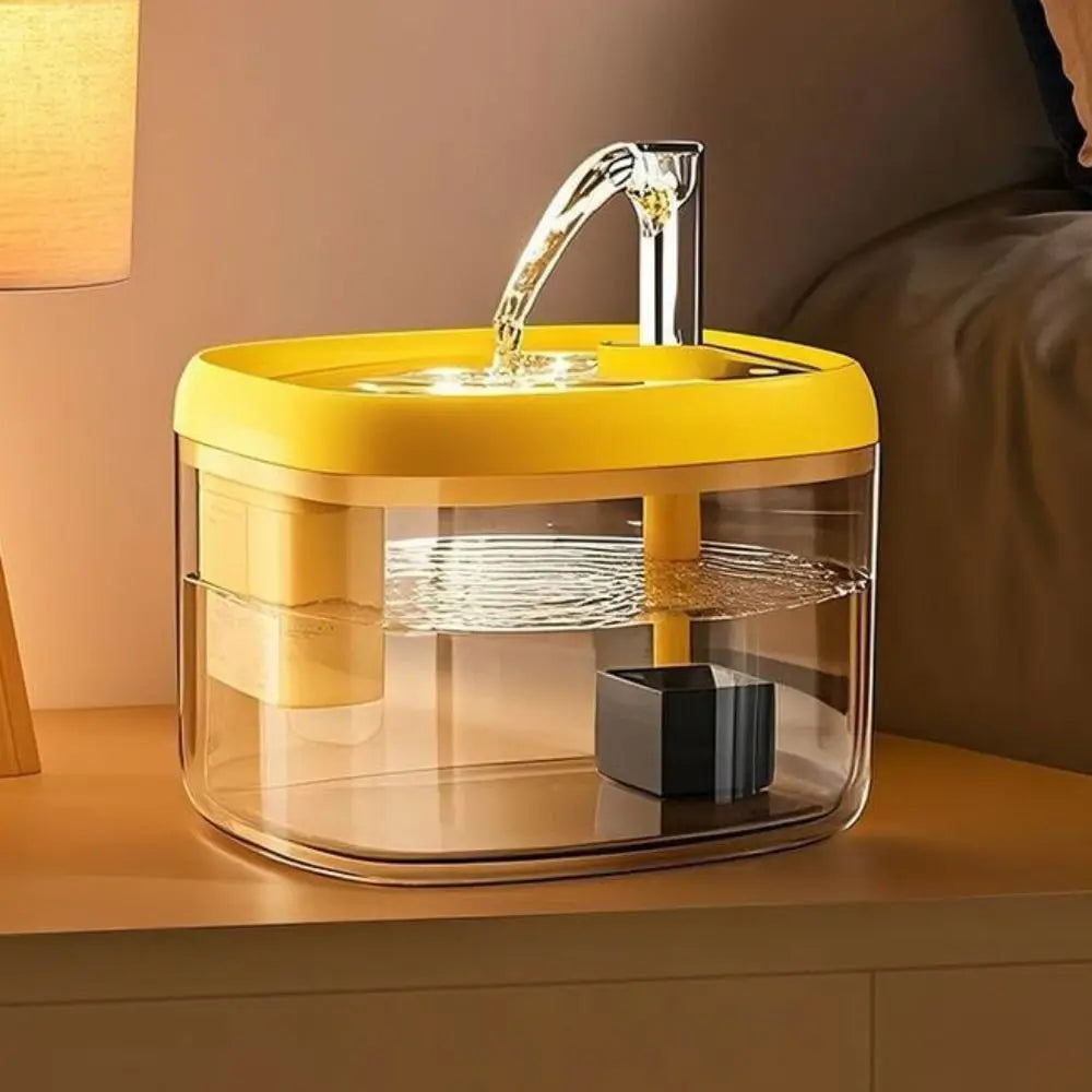A yellow and clear glass cat drinking fountain with a water spout is placed on a wooden surface.