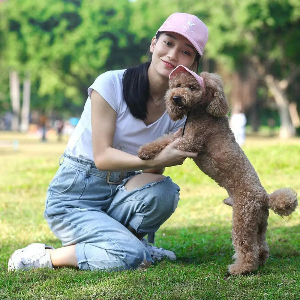 A young woman is sitting on the grass with a fluffy brown dog, both wearing pink baseball caps.