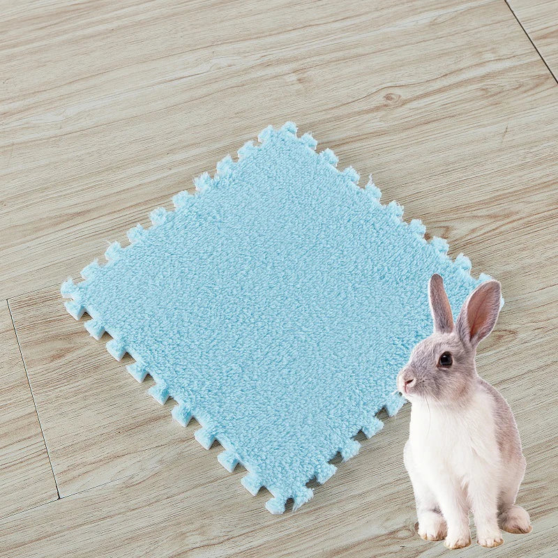 A small gray rabbit sits next to a light blue, textured fleece pet warming mat on a wooden floor.