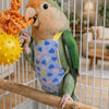 A green parrot wearing a blue patterned flight suit and holding an orange toy inside a wire cage.