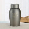A matte gray stainless steel pet memorial storage box with a black lid sitting on a light surface.