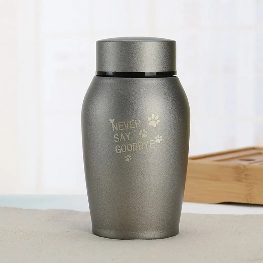 A matte gray stainless steel pet memorial storage box with a black lid sitting on a light surface.