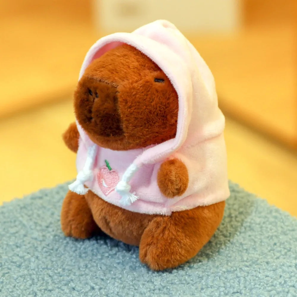 A brown capybara plush doll wearing a pink hoodie with white trim and small wings, sitting on a blue textured surface.