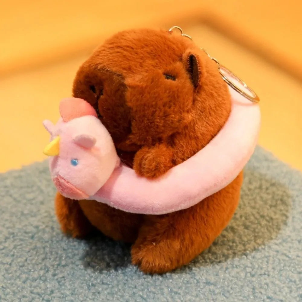 A brown capybara plush doll wearing a pink hoodie with wings, resting on a blue fabric surface.