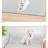 A fluffy white dog is lying on a gray quilted washable cooling mat placed on a wooden floor and a sofa.