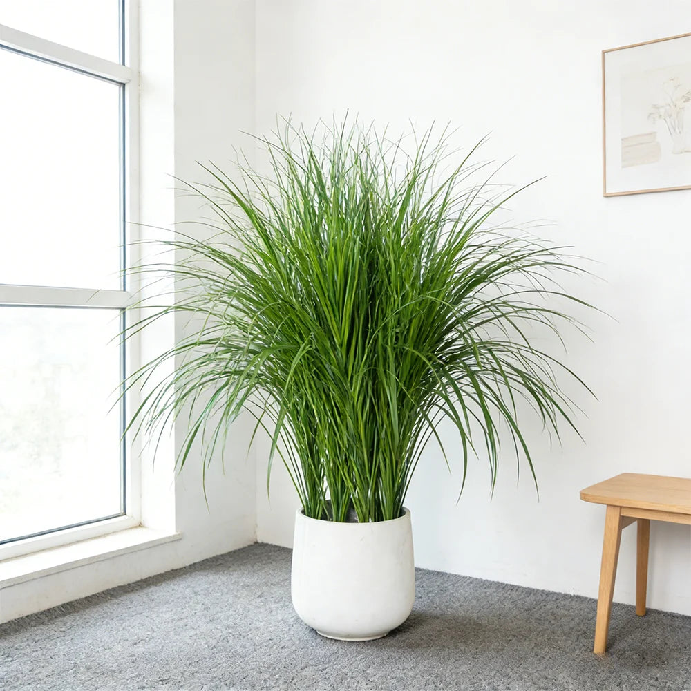 A tall potted artificial reed grass bouquet in a white ceramic pot on a gray floor next to a window.
