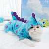 A white cat wearing a blue dinosaur fleece hoodie with purple accents, lying on a white surface.