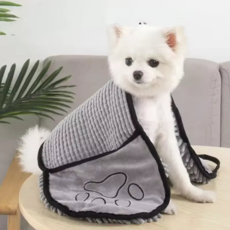 A white dog wearing a gray pet bath towel with black trim sitting on a wooden surface.