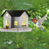 A solar outdoor bird feeder lantern shaped like a small house, with a bird flying near it in a green outdoor setting.