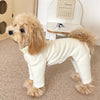 A light beige dog wearing a white soft stretch pajama jumpsuit, sitting on a carpet.